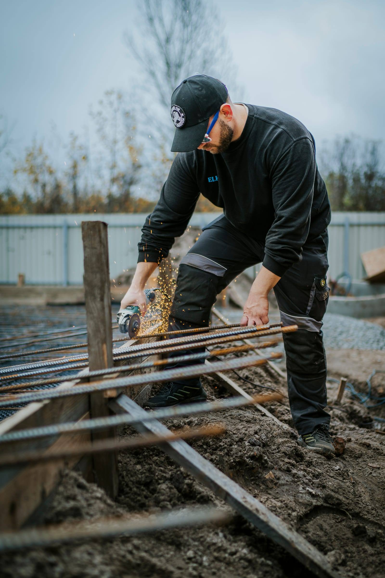 A volunteer cuts wooden stakes at a building site beside a metal fence